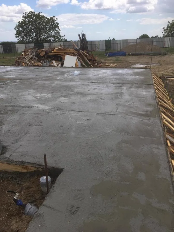 The freshly poured concrete base of the second building, timber stacked behind it.
