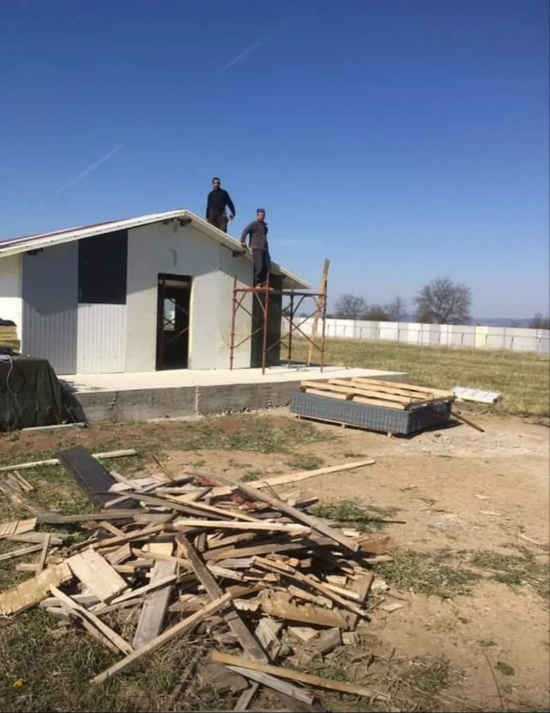 Two helpers roofing the building, timber piled in front.